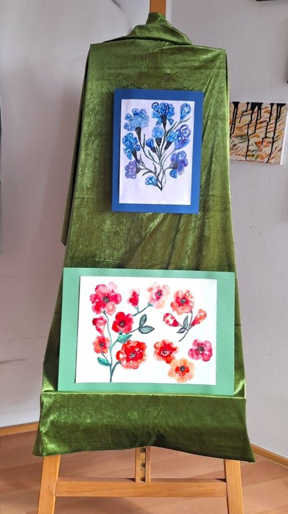 Zwei farbenfrohe Blumen‑Aquarelle auf einer Staffelei mit grünem Samt, gerahmt in Blau und Grün – stilvolle Präsentation botanischer Malerei.