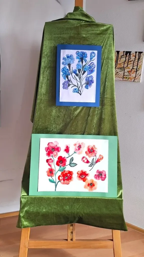 Zwei farbenfrohe Blumen‑Aquarelle auf einer Staffelei mit grünem Samt, gerahmt in Blau und Grün – stilvolle Präsentation botanischer Malerei.