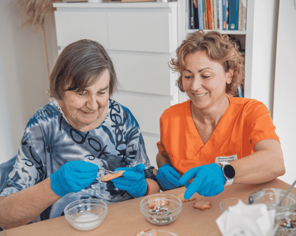Eine ältere Frau und eine Mitarbeiterin sitzen gemeinsam an einem Tisch und verzieren kleine Plätzchen. Beide tragen blaue Einmalhandschuhe. Auf dem Tisch stehen Schüsseln mit Zutaten und bunten Streuseln.