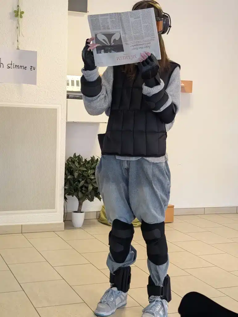 Eine Schülerin trägt den Alterssimulationsanzug GERT mit Gewichten, Handschuhen und Simulationsbrille und hält eine Zeitung vor ihr Gesicht.