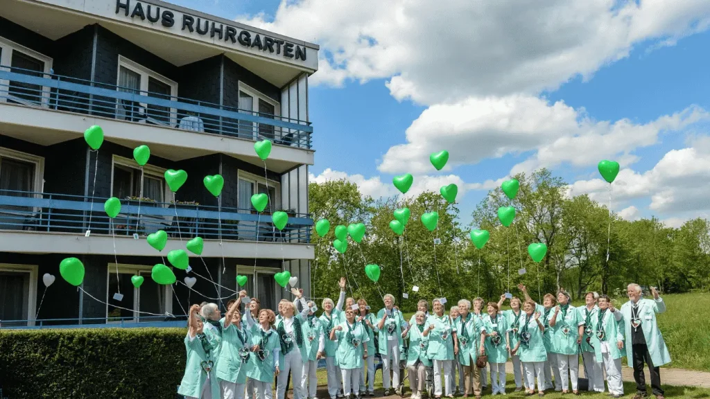 Eine große Gruppe der Grünen Damen und Herren steht vor dem Haus Ruhrgarten. Die Ehrenamtlichen tragen einheitliche mintfarbene Kleidung und halten grüne Herzluftballons in den Händen. Im Hintergrund sind das Gebäude des Hauses Ruhrgarten und eine grüne Parklandschaft unter blauem Himmel zu sehen.