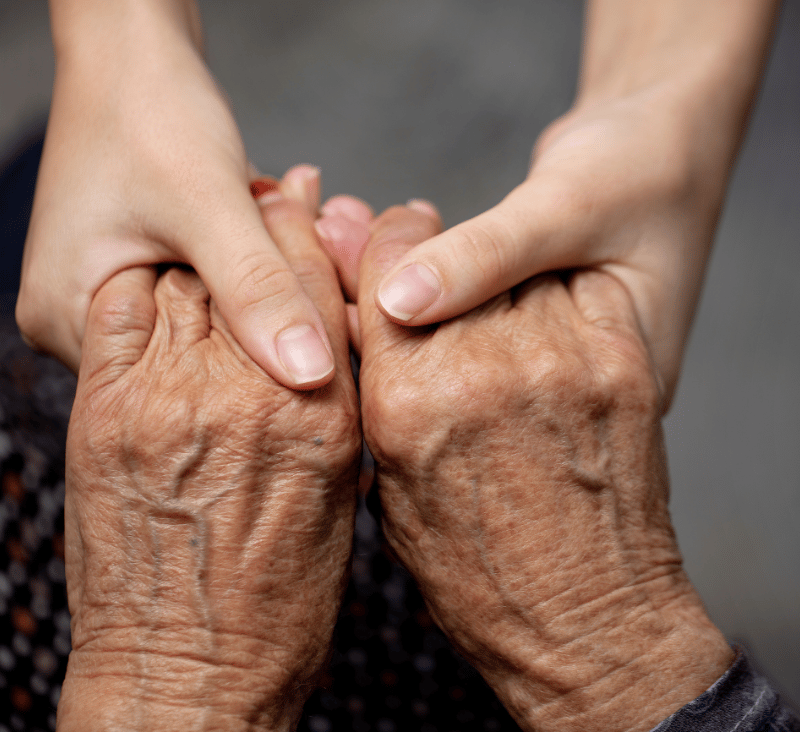 Nahaufnahme zweier Hände: Eine jüngere Hand hält und berührt behutsam die Hände einer älteren Person. Das Bild vermittelt Nähe, Fürsorge und menschliche Verbundenheit.