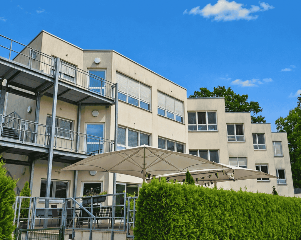 Außenansicht des Hauses Ruhrblick mit hell verputztem mehrstöckigem Gebäude, Balkonen und großen Fenstern. Im Vordergrund befinden sich eine begrünte Terrasse mit Sonnenschirmen und Hecken. Darüber ist ein strahlend blauer Himmel zu sehen.