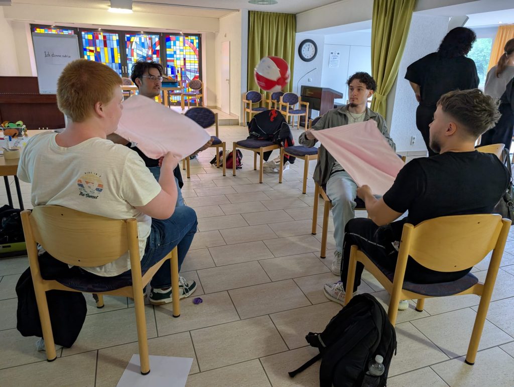 Mehrere Schüler:innen sitzen im Kreis und bewegen gemeinsam ein großes Tuch mit einem Ball während einer Aktivierungsübung im Haus Ruhrgarten.