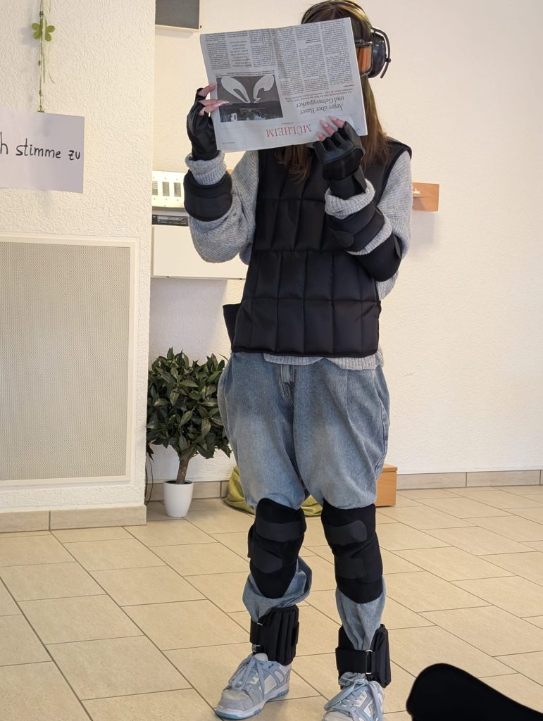 Eine Schülerin trägt den Alterssimulationsanzug GERT mit Gewichten, Handschuhen und Simulationsbrille und hält eine Zeitung vor ihr Gesicht.