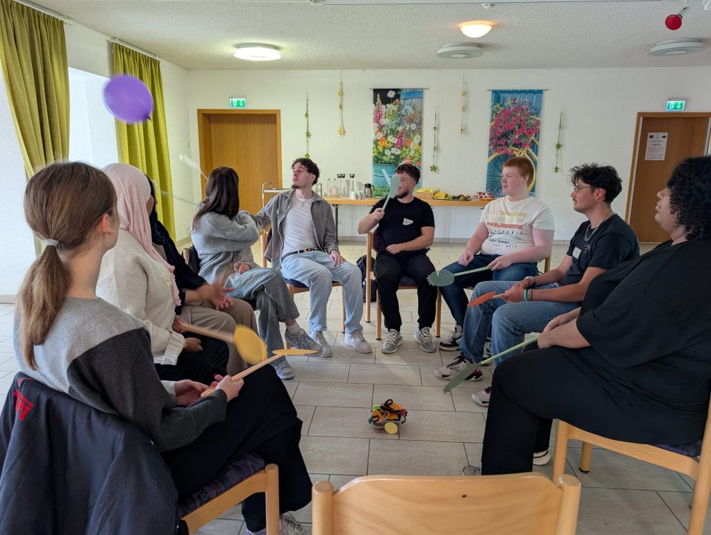 Schüler:innen des Berufskollegs Stadtmitte sitzen im Kreis und führen gemeinsam mit einem Luftballon und bunten Fliegenklatschen eine Bewegungsübung im Haus Ruhrgarten durch.