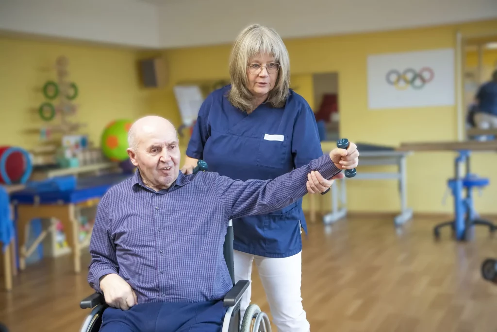Therapeutin unterstützt den linken ausgestreckten Arm eines Bewohners im Rollstuhl, der eine kleine Hantel in der Hand hält.