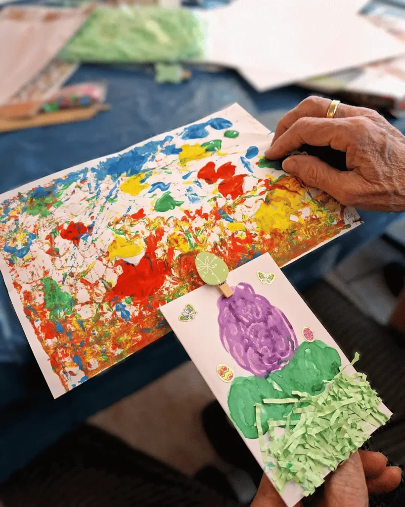 Nahaufnahme einer Person beim kreativen Gestalten. Auf einem Tisch liegen farbenfrohe, mit verschiedenen Farben bemalte Bilder. Im Vordergrund ist ein selbst gestaltetes Bild mit einer großen lila Blüte und grünen Blättern zu sehen.