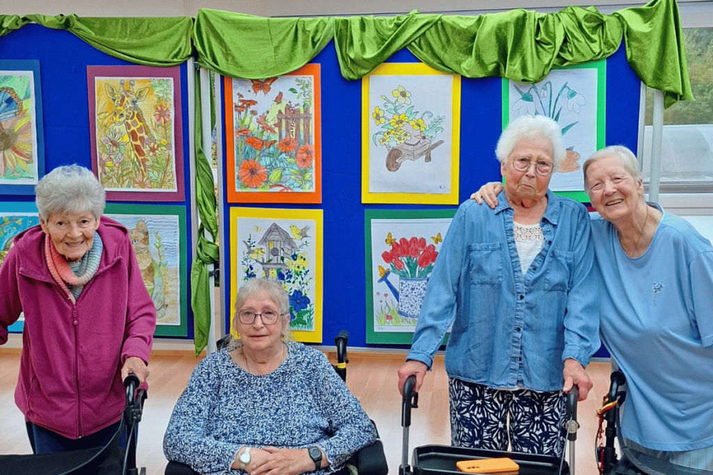 Vier Bewohner:innen des Haus Ruhrblick in Mülheim an der Ruhr stehen vor einer Ausstellung ihrer selbst geschaffenen Kunstwerke aus der Maltherapie.