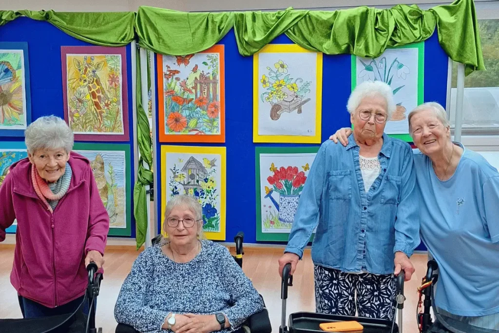 Vier Bewohner:innen des Haus Ruhrblick in Mülheim an der Ruhr stehen vor einer Ausstellung ihrer selbst geschaffenen Kunstwerke aus der Maltherapie.