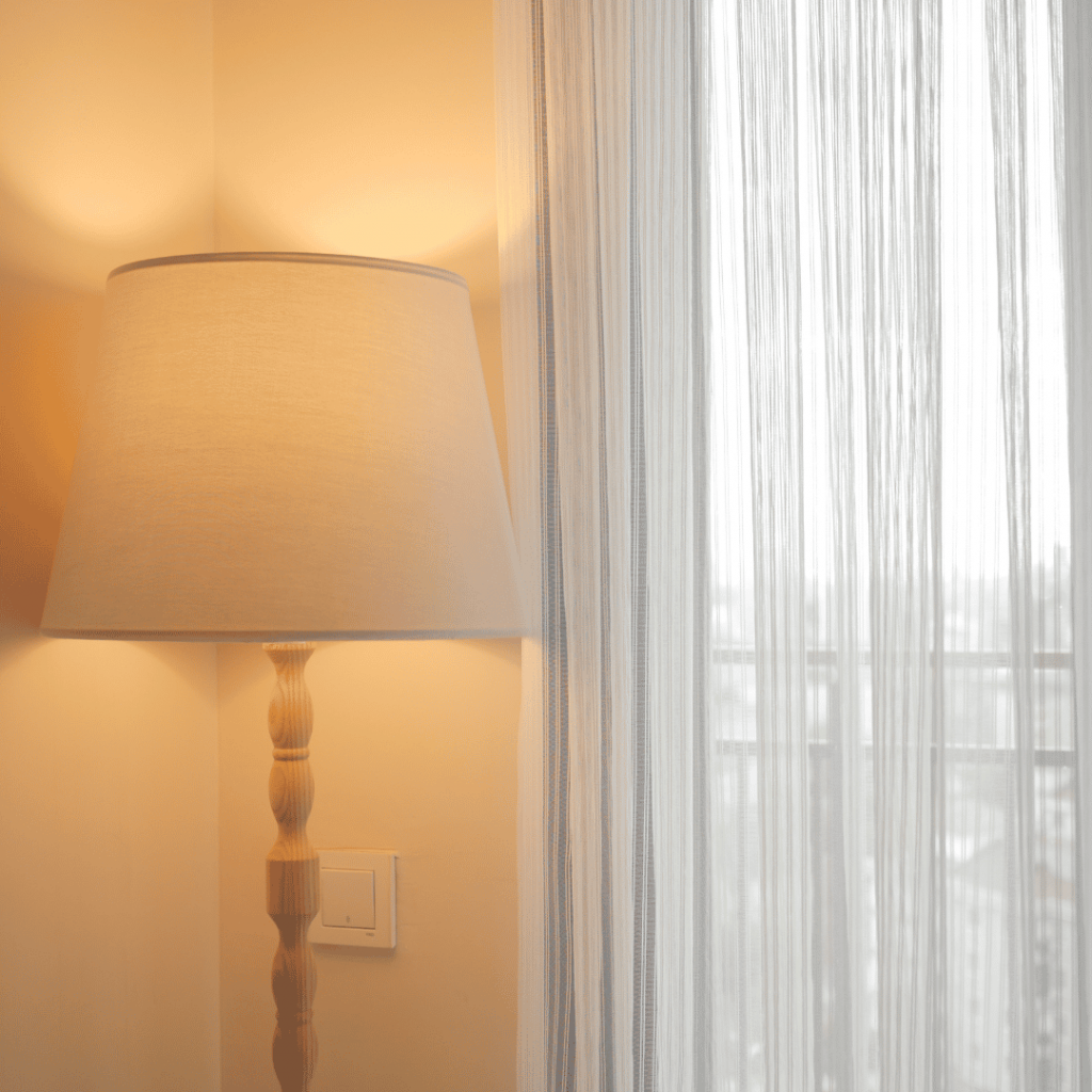 Eine eingeschaltete Stehlampe mit warmem Licht steht neben einem Fenster mit weißen, lichtdurchlässigen Vorhängen.