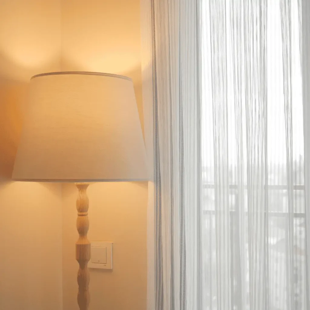 Eine eingeschaltete Stehlampe mit warmem Licht steht neben einem Fenster mit weißen, lichtdurchlässigen Vorhängen.