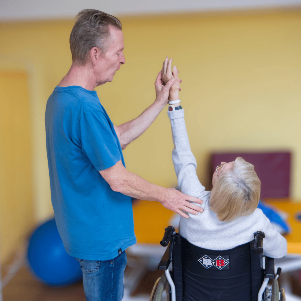 Ein Therapeut unterstützt eine Bewohnerin im Rollstuhl bei einer Bewegungsübung. Die Bewohnerin streckt einen Arm nach oben, während der Mitarbeiter ihre Bewegung begleitet. Im Hintergrund sind Gymnastikbälle und weitere Sportgeräte zu sehen.