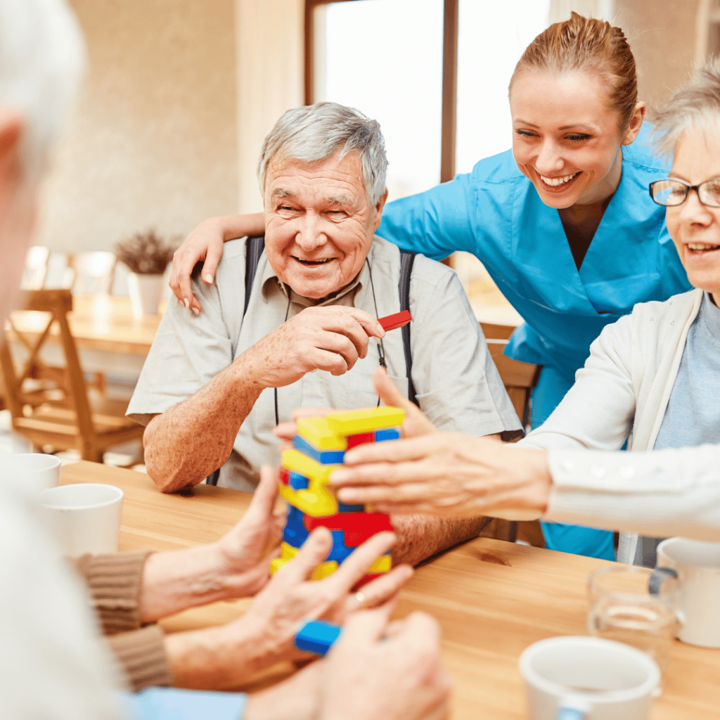 Ein älterer Mann sitzt gemeinsam mit einer Pflegekraft und einer weiteren Person an einem Tisch und beschäftigt sich mit einem bunten Geschicklichkeitsspiel. Die Pflegekraft lächelt ihm zu und begleitet die gemeinsame Aktivität.