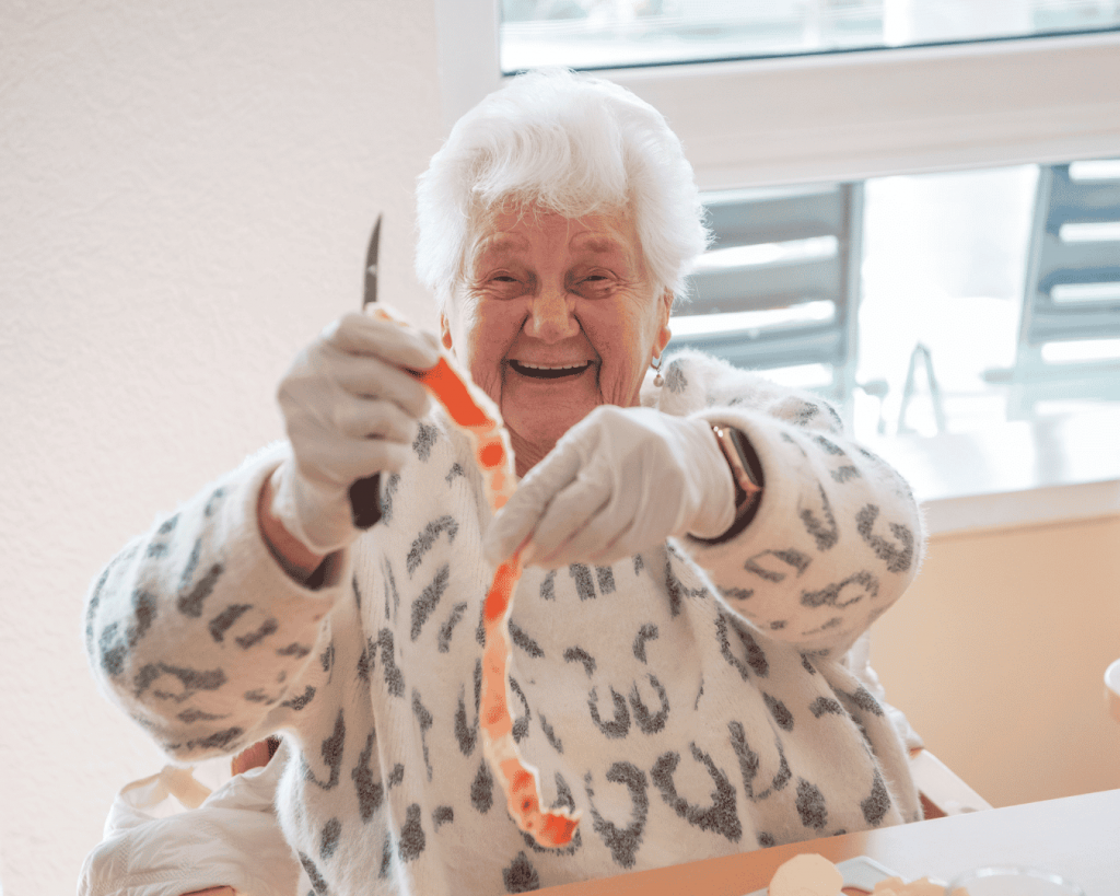 Eine ältere Frau sitzt bei einer kreativen und hauswirtschaftlichen Beschäftigung am Tisch. Sie hält ein Messer in der Hand und hat einen Apfel geschält; die Apfelschale hält sie in der anderen Hand. Sie trägt einen gemusterten Pullover und lacht sehr herzlich.