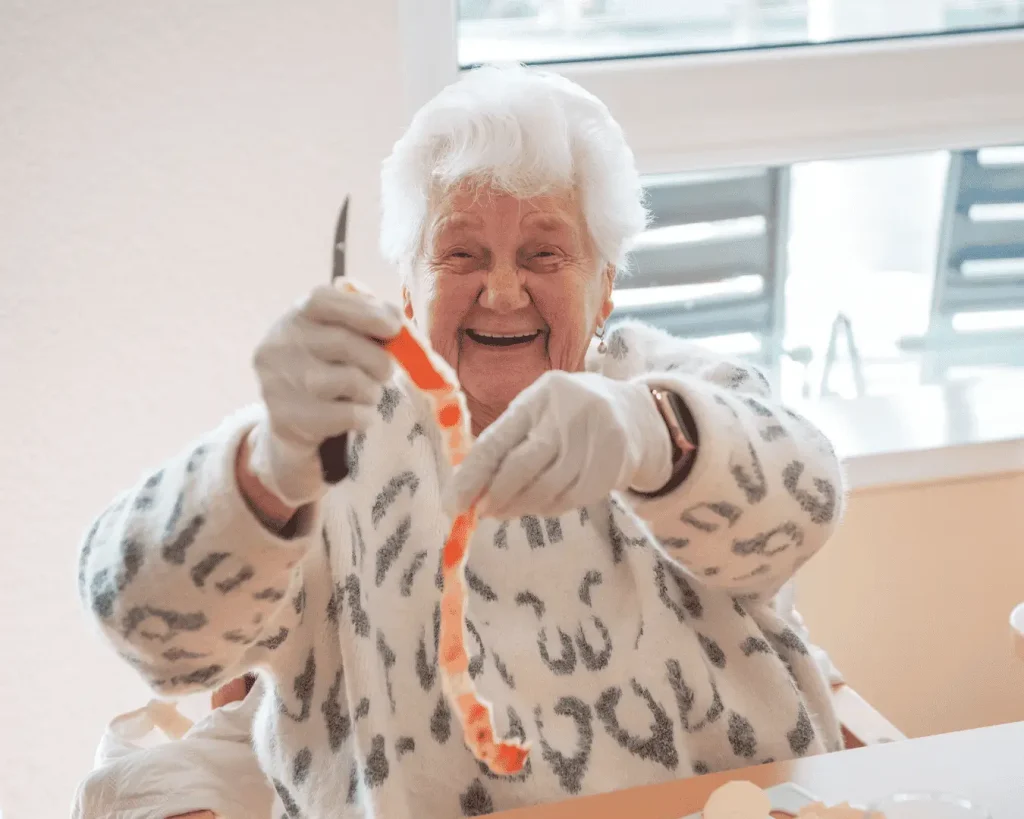 Eine ältere Frau sitzt bei einer kreativen und hauswirtschaftlichen Beschäftigung am Tisch. Sie hält ein Messer in der Hand und hat einen Apfel geschält; die Apfelschale hält sie in der anderen Hand. Sie trägt einen gemusterten Pullover und lacht sehr herzlich.