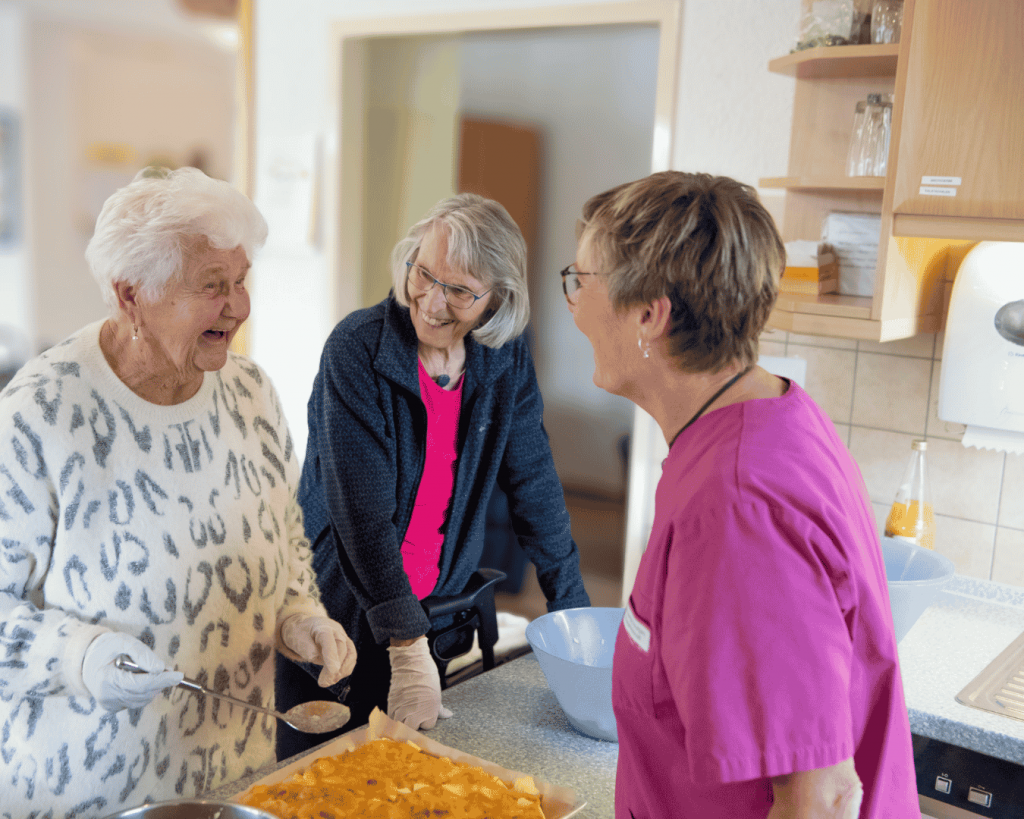 Zwei ältere Frauen bereiten gemeinsam mit einer Betreuungskraft in einer Küche einen Kuchen oder Teig zu. Eine Frau hält einen Löffel über eine Schüssel, während die andere den Inhalt einer Backform betrachtet. Die drei Frauen lachen und unterhalten sich.