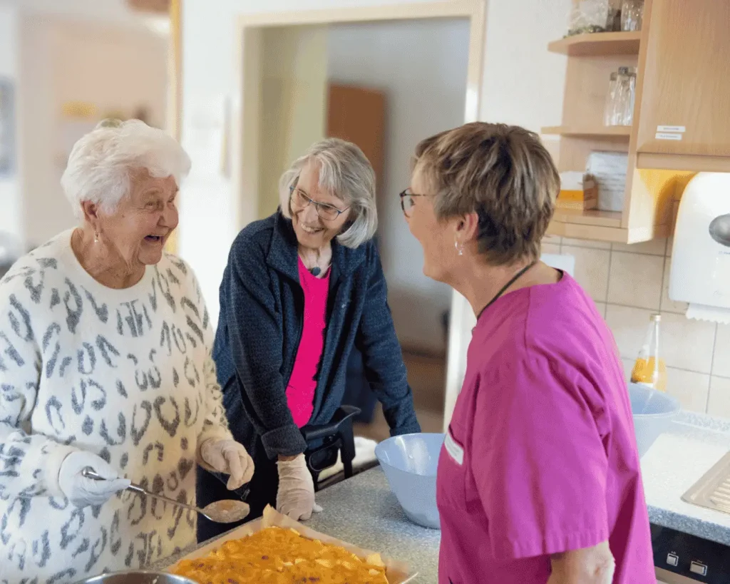 Zwei ältere Frauen bereiten gemeinsam mit einer Betreuungskraft in einer Küche einen Kuchen oder Teig zu. Eine Frau hält einen Löffel über eine Schüssel, während die andere den Inhalt einer Backform betrachtet. Die drei Frauen lachen und unterhalten sich.